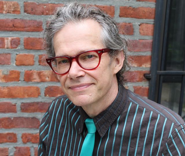 Image of Mike Putnam. He's a white man with red glasses, wearing a teal colored tie and a shirt with thin stripes of the same color. Mike looks friendly and confident. He is looking directly into the camera. There's a brick wall behind him. 