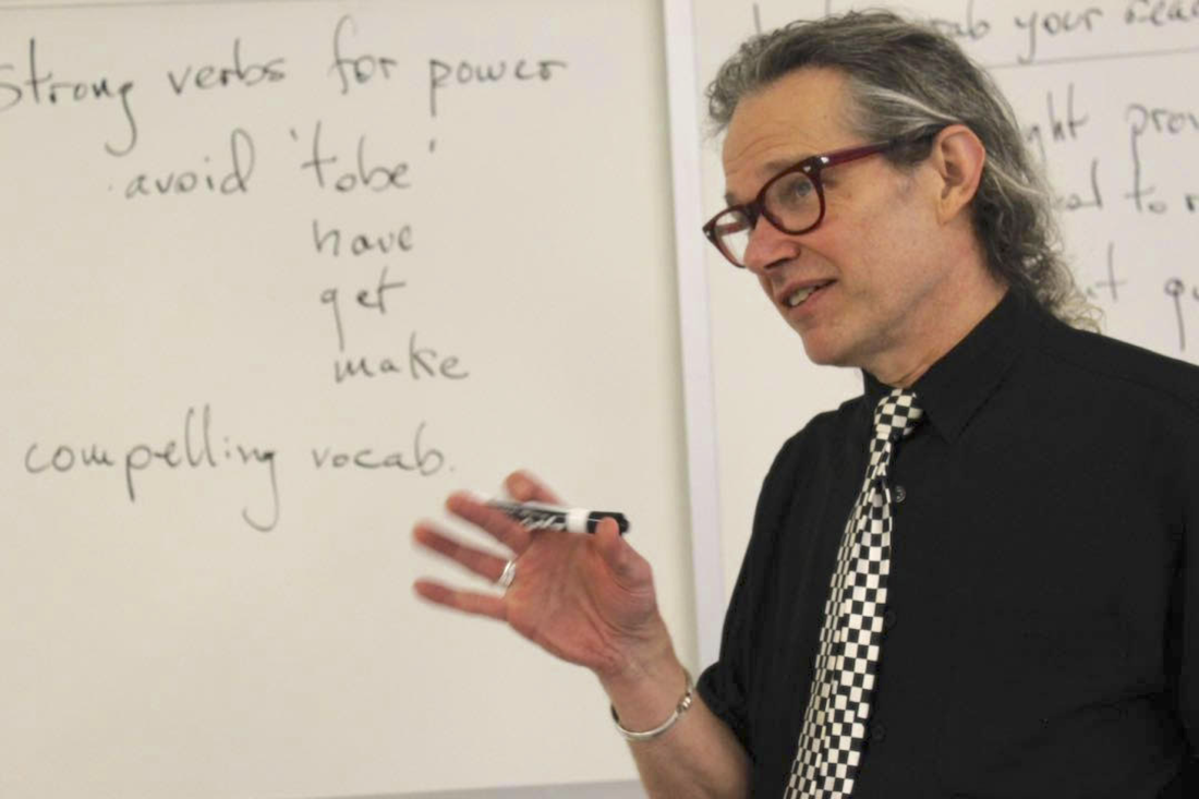 Mike Putnam, in front of a white board with a marker in his hand. He's making a presentation or teaching. His hand is moving upward as if he's engaged with participants and making a point. His focus appears to be on someone to our left. He's wearing a black shirt, with a funky black & white tie, and big red glasses. 