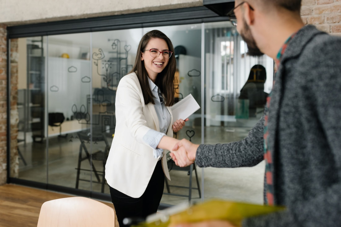 Image of woman greeting a man, she has a big smile and strong handshake. Focus in on her, we see only the back of his head. She's wearing a casual white suit jacket and is holding some papers. He is in a grey sweater. 