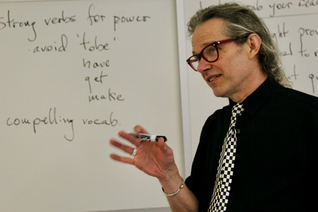 Mike teaching a writing class, glasses on, black shirt with checker board tie, marker in hand.
